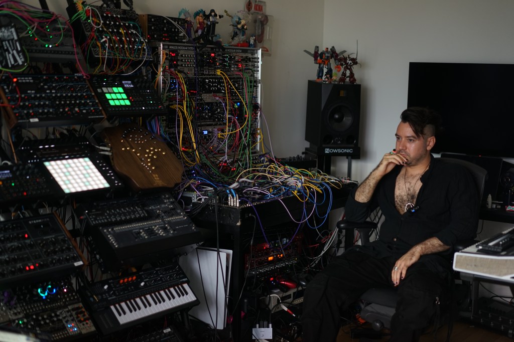 A musician sitting in a studio surrounded by various synthesizers and electronic equipment, deep in thought.